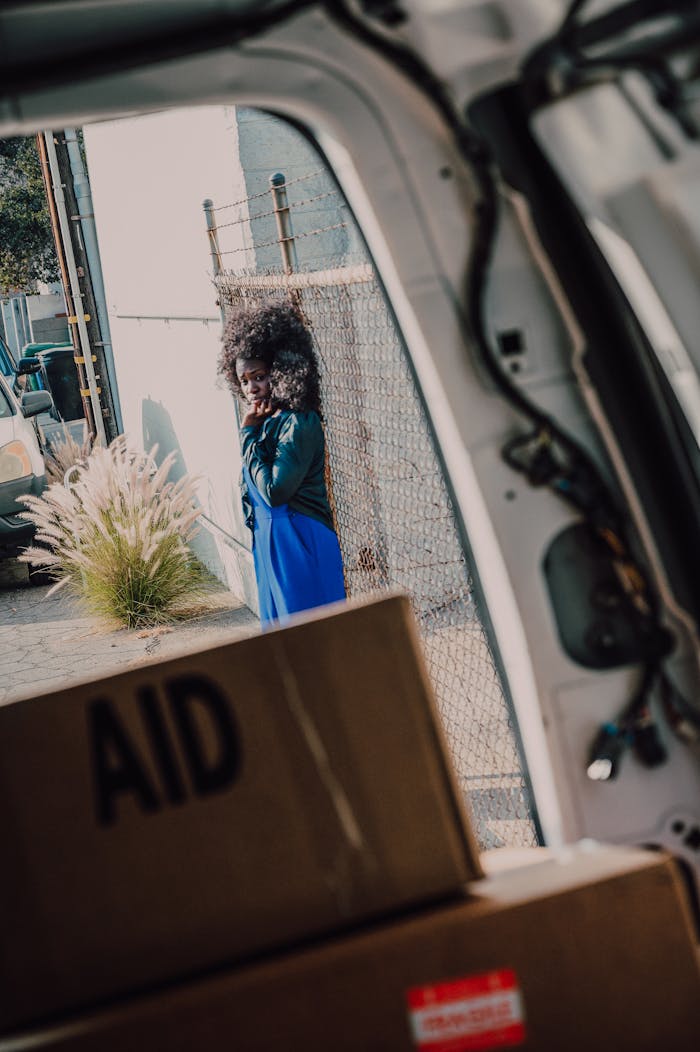 Woman in blue dress beside aid boxes viewed from a vehicle.